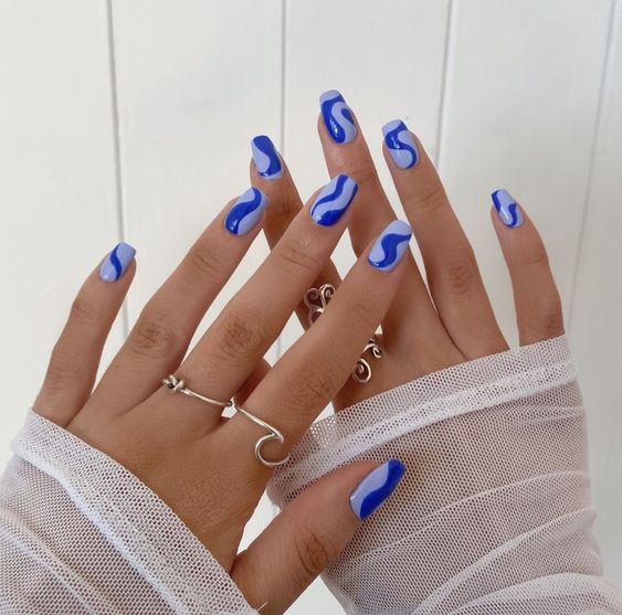 Mãos de pele branca, com anéis prata e unha quadrada azul-claro com linhas fluidas 