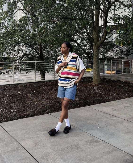 Mulher jovem, magra e negra, usando o cabelo preso, uma camiseta branca oversized, colete colorido em sobreposição, bermuda jeans e mocassim