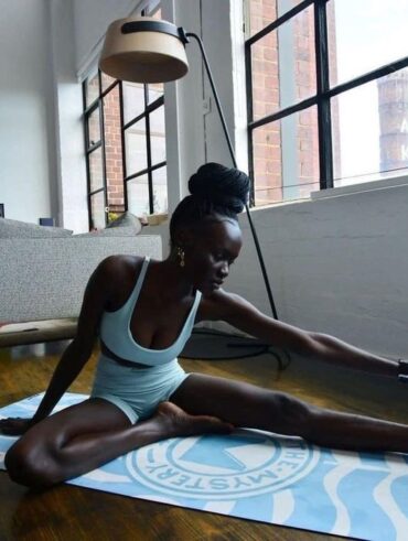 Mulher jovem e magra com pele escura. Ela está sentada em um tapete de exercício fazendo alongamento.