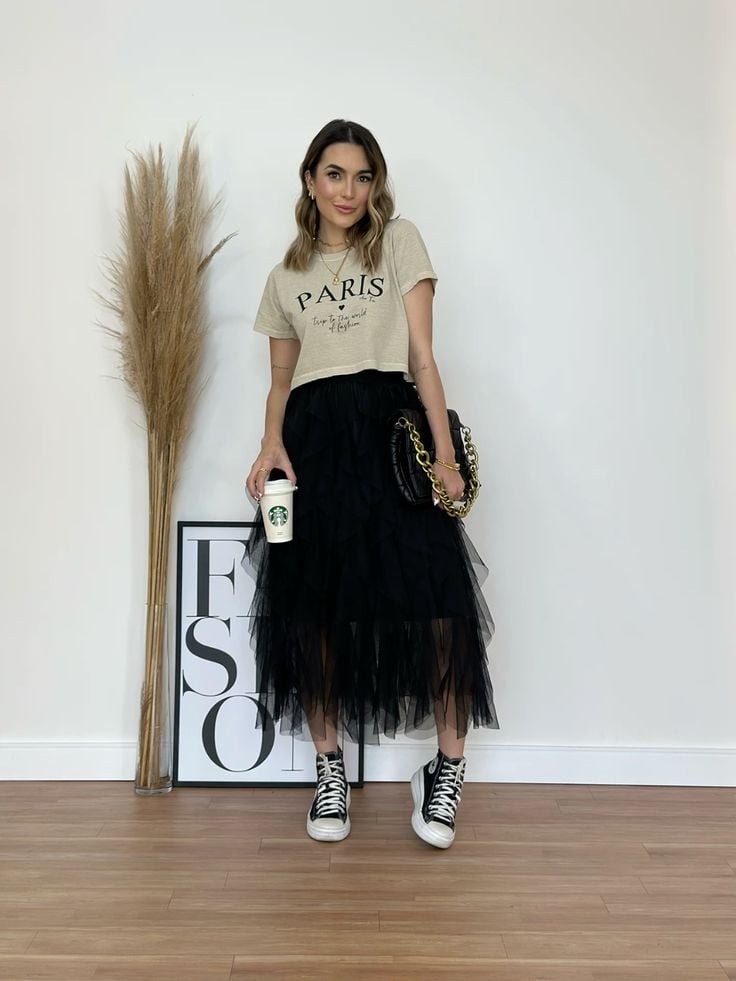 Mulher loira com cabelo curto, usando camiseta bege, saia de tule preta, All Star de cano alto e segurando uma bolsa e um copo de café