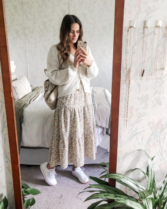 Mulher jovem e branca, usando vestido longo estampado com flores e no modelo três marias, tênis branco e uma mochila