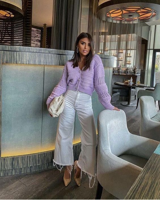Mulher jovem, magra e de pele clara. Ela veste cardigan lilás, calça jeans branca e sapato dourado.