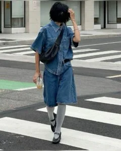 Mulher jovem, magra e de pele clara. Ela usa conjunto jeans, meia cinza e sapato preto.