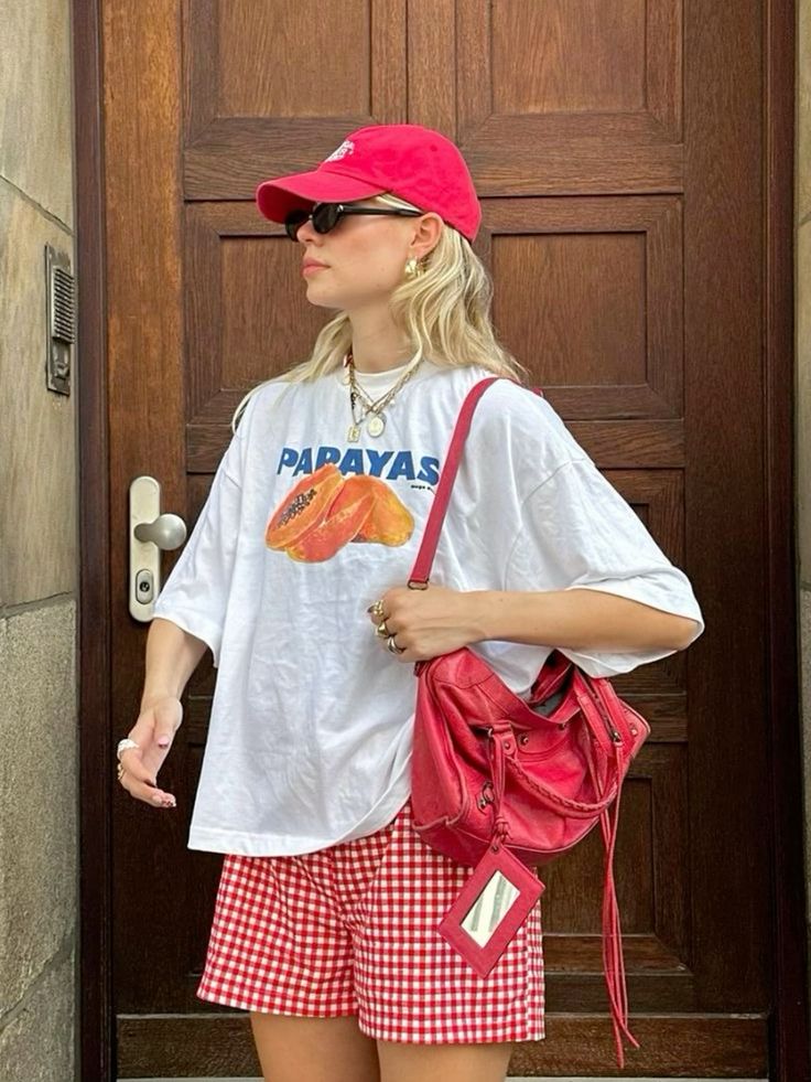 Mulher jovem, magra e de pele clara com cabelos loiros. Ela usa camiseta com estampa de fruta, bermuda e boné.