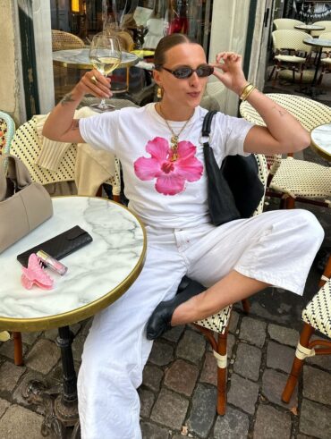 Mulher jovem e de pele clara. Ela está com calça branca, sapatilha preta e camiseta estampada de flor rosa.