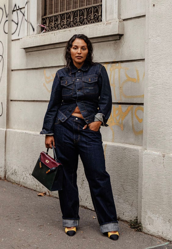 Mulher jovem mid size com cabelo escuro. Ela veste jaqueta e calça jeans.
