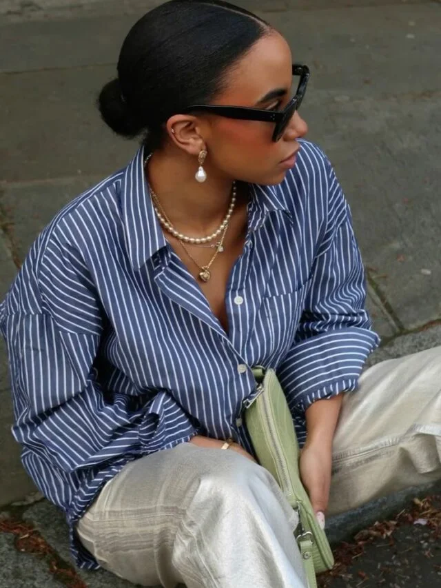 Mulher jovem e negra, com cabelo preso em coque, usando óculos de sol, camisa azul listrada, colar de perolas e segurando uma bolsa pequena em cor verde