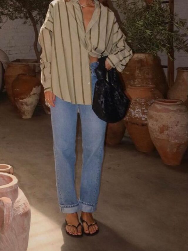 Mulher usando óculos de sol, camisa bege listrada em marrom, calça jeans, chinelo e segurando uma bolsa.