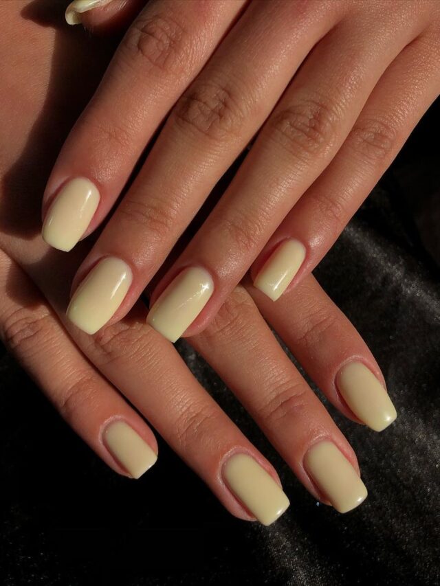 Mão de pele branca, com unha média e quadrada, com esmalte amarelo-manteiga em tom pastel