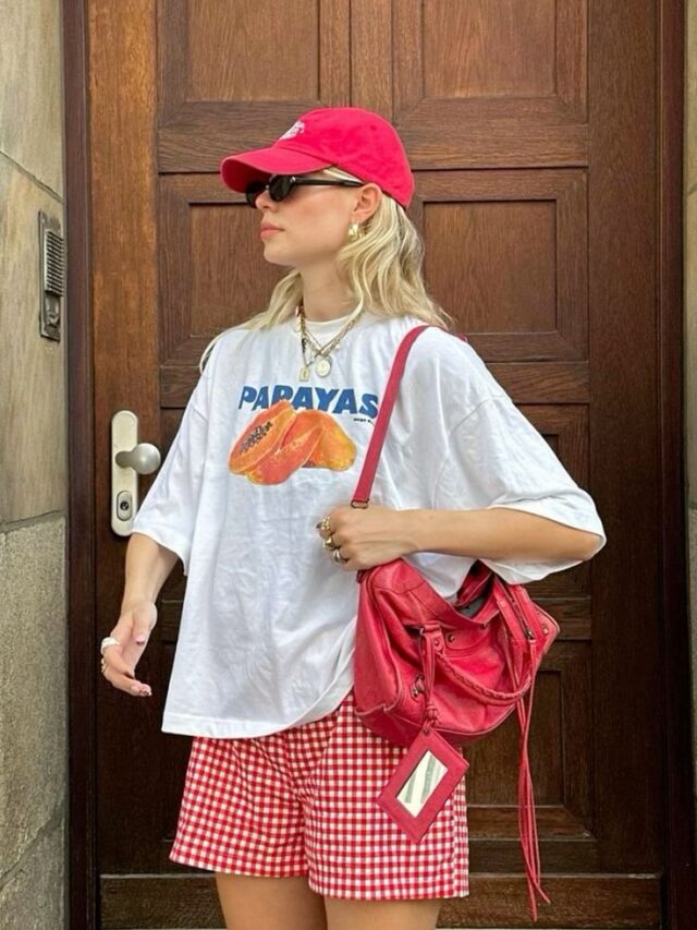 Mulher jovem, magra e de pele clara com cabelos loiros. Ela usa camiseta com estampa de fruta, bermuda e boné.