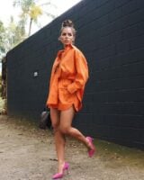 Mulher jovem, usando coque no cabelo, conjunto de camisa e short laranja com sapato de salto em cor rosa