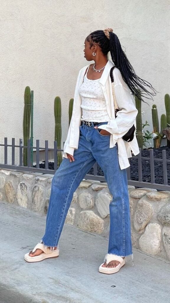 Mulher jovem, magra e negra, usando camisa branca com blusa na mesma tonalidade, cabelo preso em rabo de cavalo, calça jeans com cinto e chinelo.