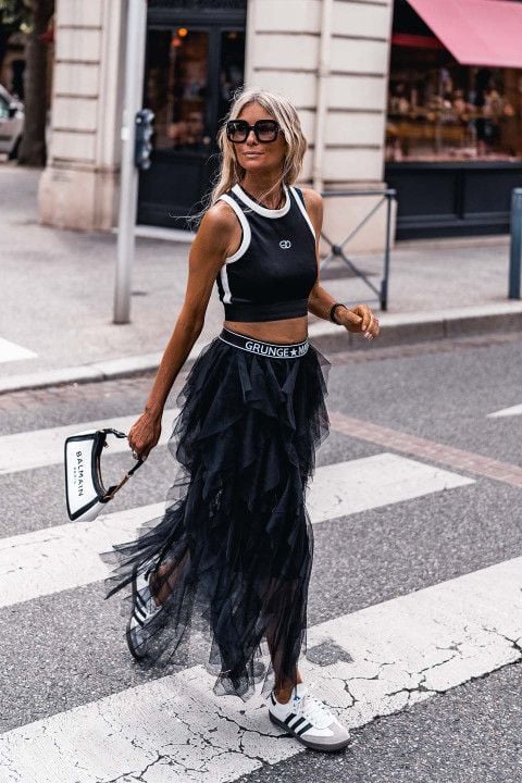 Mulher lira, usando óculos de sol, cropped de manga curta em cor preta, saia de tule preta, Adidas Samba e segurando uma bolsa