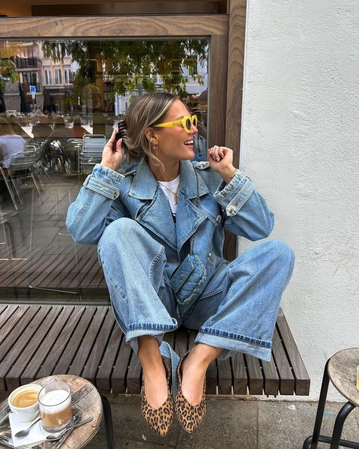 Mulher jovem e magra de pele clara. Ela está sentada em uma cadeira com os pés para cima e usa sapatilha de oncinha e look all jeans.