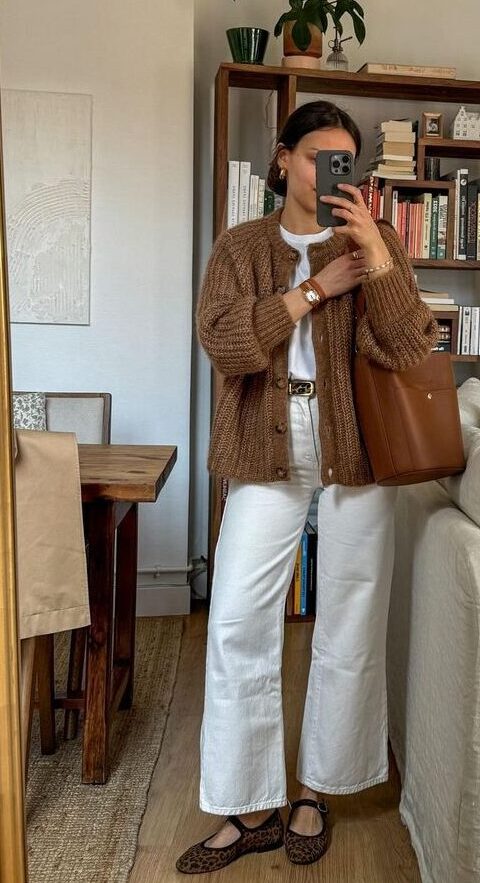 Mulher jovem e magra em frente ao espelho. Ela usa look branco com cardigan marrom e calçado estampado.