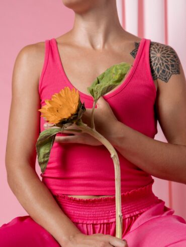 Tronco de uma mulher em campanha contra o câncer de mama; ela usa regata e calça rosa e está segurando uma flor.