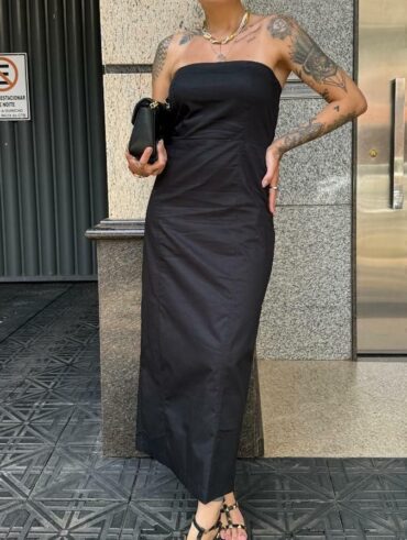 Mulher jovem, branca e com cabelo escuro preso em coque, com uma mão na cintura, usando vestido longo sem alça, sandália e segurando uma bolsa preta.