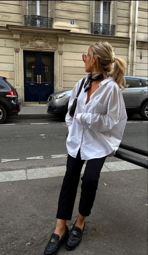 Mulher jovem, magra e loira, usando o cabelo preso, lenço no cabelo, camisa branca no estilo oversized, calça reta na cor preta, bolsa de ombro e mocassim.