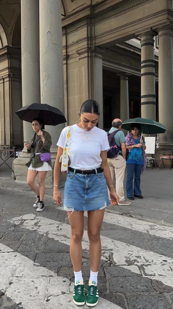 Mulher branca e com cabelo moreno preso em coque, usando camiseta branca, saia jeans com cinto preto, meia longa com tênis verde e bolsa de ombro
