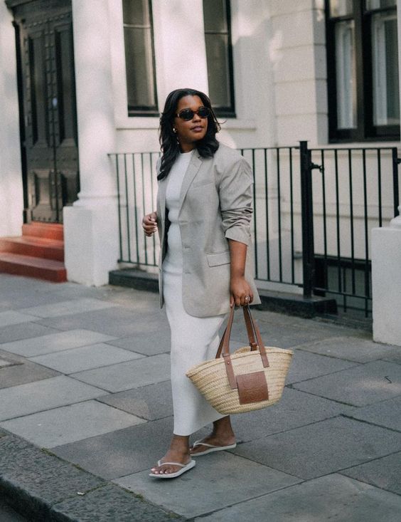 Mulher jovem e negra, usando óculos de sol, blazer cinza e vestido branco com chinelo e uma bolsa de palha