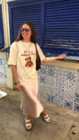 Mulher jovem e branca, usando óculos de sol, camiseta na cor bege estampada, saia longa de cetim, bolsa de ombro na cor marrom e papete