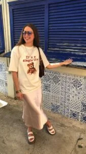 Mulher jovem e branca, usando óculos de sol, camiseta na cor bege estampada, saia longa de cetim, bolsa de ombro na cor marrom e papete