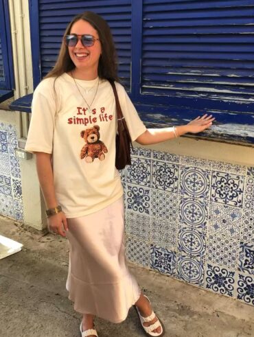 Mulher jovem e branca, usando óculos de sol, camiseta na cor bege estampada, saia longa de cetim, bolsa de ombro na cor marrom e papete