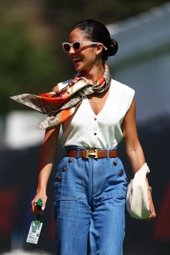 Mulher branca e magra, com cabelo preso em coque, usando óculos de sol, lenço no pescoço, blusa regata de botões na cor branca e calça jeans de cintura alta com cinto marrom.