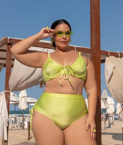 Mulher plus size, branca e jovem, usando cabelo preso em coque, óculos de sol e biquíni meia taça na cor verde.