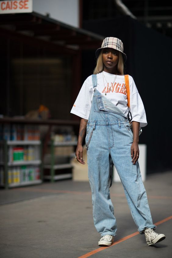 Mulher jovem e negra, usando bucket xadrez, camiseta oversized, jardineira jeans e All Star.