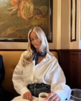 Mulher jovem e branca, com cabelo loira, sentada em mesa de café, usando lenço no pescoço na cor azul, camisa branca no estilo oversized e calça jeans.