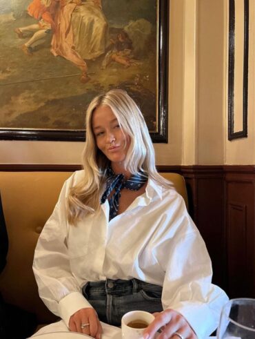 Mulher jovem e branca, com cabelo loira, sentada em mesa de café, usando lenço no pescoço na cor azul, camisa branca no estilo oversized e calça jeans.