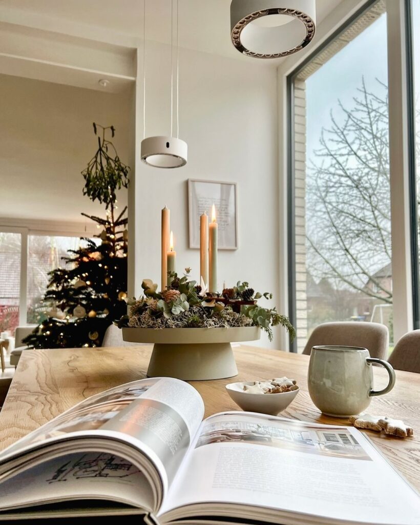 Mesa de jantar com centro de mesa de Natal, com pinhas e velas. Na mesa, estão ainda um livro e uma xícara