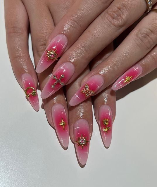 Mãos de pele branca, com unha aura rosa amendoada com charms em dourado 