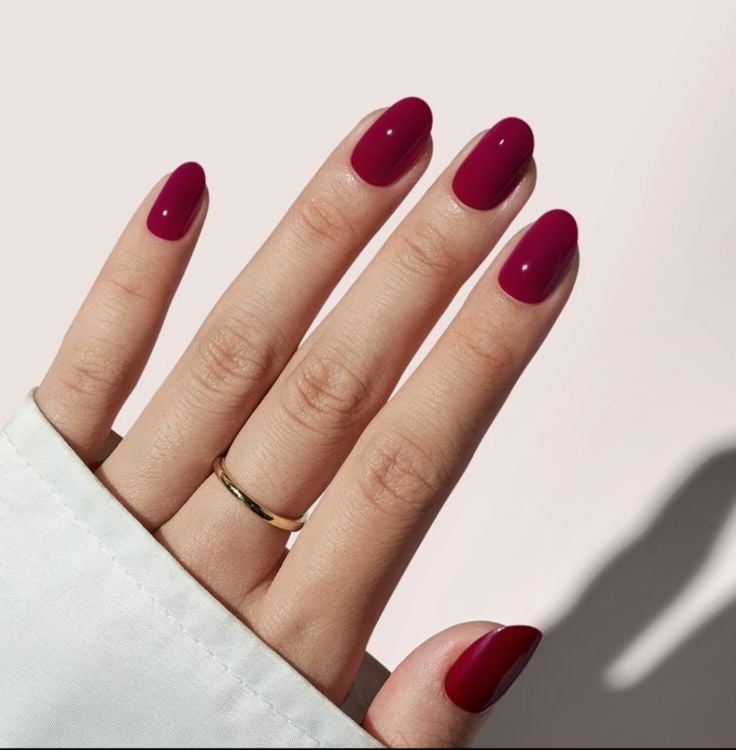 Mão de pele branca, com anel em dourado e unha curta e amendoada vermelho-cereja 