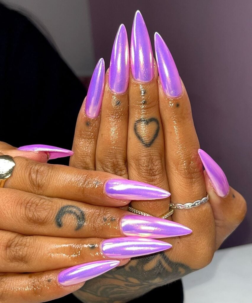 Mãos de pele preta, com tatuagens, anéis em prata e unha stiletto comprida lilás com efeito cromado