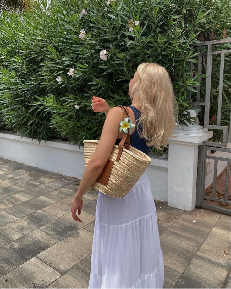 Mulher jovem, magra e de pele claro. Ela está de costas com roupas claras e usa uma bolsa de palha média nos ombros.