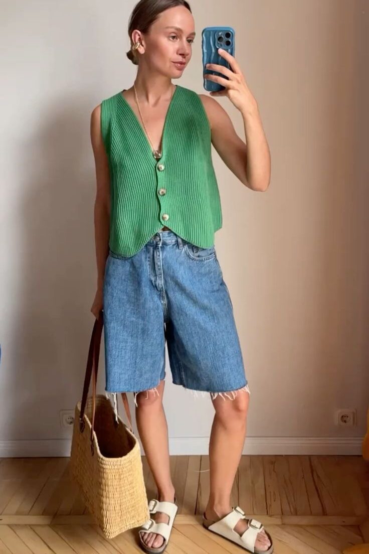 Mulher jovem e magra usando colete verde, bermuda jeans e calçado branco.