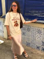 Mulher jovem e branca, usando óculos de sol, camiseta na cor bege estampada, saia longa de cetim, bolsa de ombro na cor marrom e papete