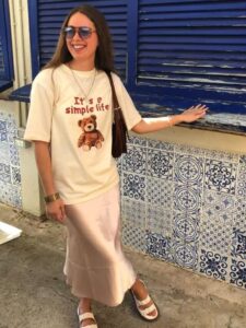 Mulher jovem e branca, usando óculos de sol, camiseta na cor bege estampada, saia longa de cetim, bolsa de ombro na cor marrom e papete