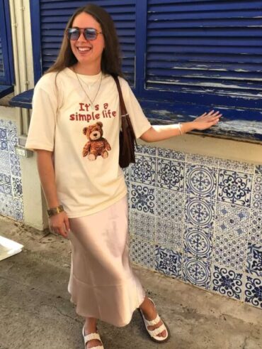 Mulher jovem e branca, usando óculos de sol, camiseta na cor bege estampada, saia longa de cetim, bolsa de ombro na cor marrom e papete