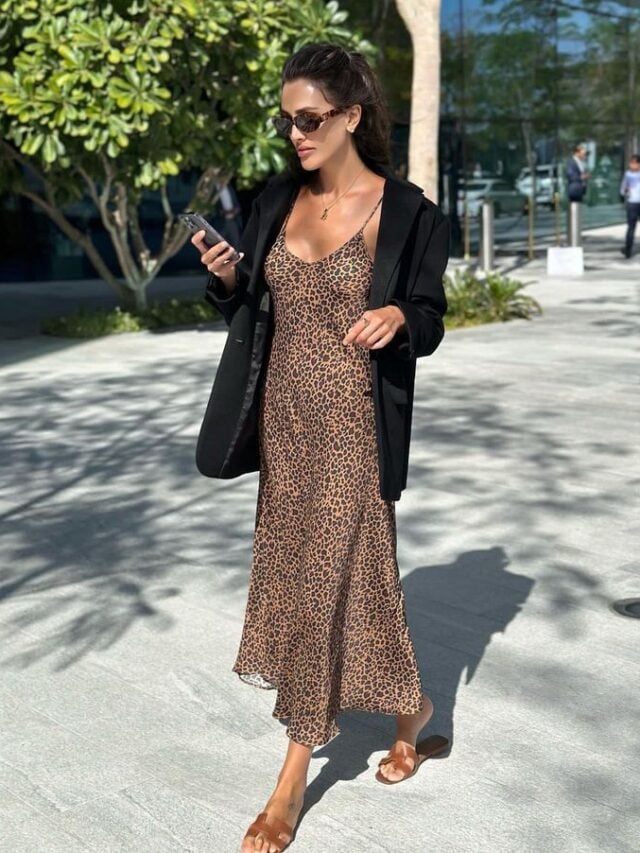 Mulher jovem e branca, segurando um celular, usando cabelo preso, óculos de sol, blazer preto, vestido longo de oncinha e rasteirinha.
