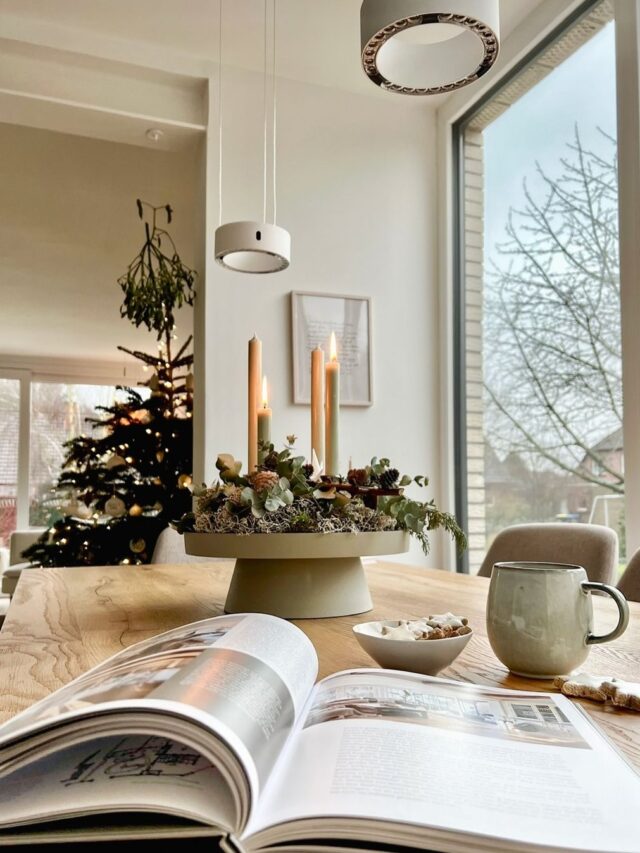 Mesa de jantar com centro de mesa de Natal, com pinhas e velas. Na mesa, estão ainda um livro e uma xícara