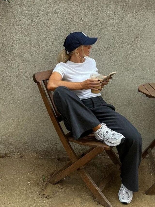 Mulher jovem e loira, sentada em uma cadeira, usando boné com cabelo preso, camiseta branca, calça de alfaiataria e New Balance 530