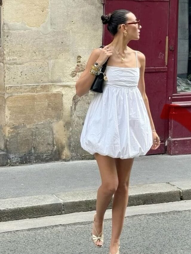 Mulher jovem e magra com pele clara. Ela está usando um vestido branco balonê de comprimento curto.
