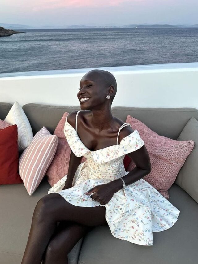 Mulher jovem e magra de pele negra. Ela usa vestido branco com flores.