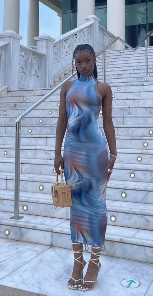 Mulher jovem e negra, usando vestido segunda pele de tule estampado em cores azuis, segurando uma bolsa pequena de palha e usando sandália de salto.
