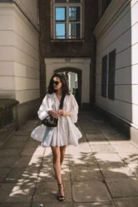 Mulher magra, branca e de cabelo curto na cor castanho escuro, usando óculos de sol, vestido branco com bolsa preta e sandália de salto.