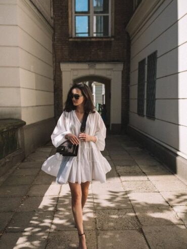 Mulher magra, branca e de cabelo curto na cor castanho escuro, usando óculos de sol, vestido branco com bolsa preta e sandália de salto.
