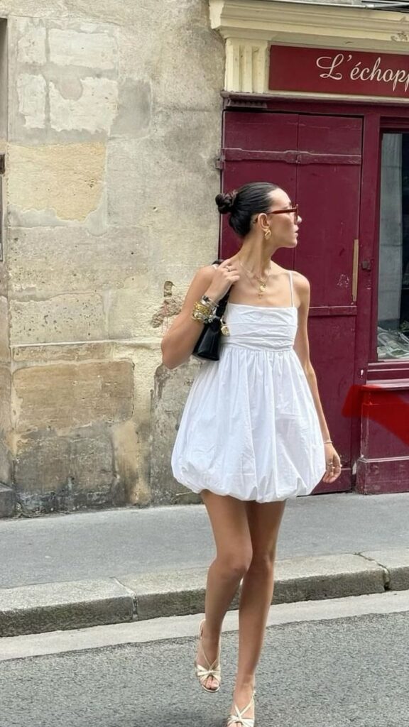 Mulher jovem e magra com pele clara. Ela está usando um vestido branco balonê de comprimento curto.